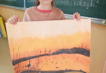 Dziewczynka stoi na tle zapisanej tablicy.Trzyma dużą kartke papieru na, której namalowany jest obraz .