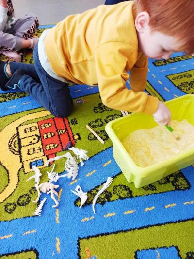 Chłopczyk szuka za pomocą pędzelka w seledynowej misce z piaskiem szkieletu dinozaurów.