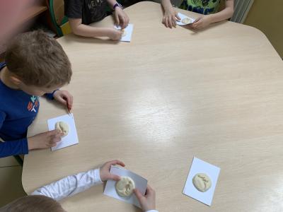 Dzieci siedzą przy stole i robią odcisk małej zabawki dinozaura w kulce masy solnej położonej na karteczce