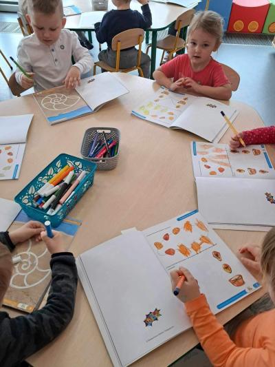 Dzieci siedzą przy stolikach w sali przedszkolnej i wykonują ćwiczenia w kartach pracy związane z przerabianą tematyką
