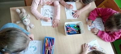 Dzieci siedzą przy stoliku i malują. Kolorowanki przedstawiają różne dinozaury