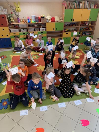 Dzieci siedzą w sali przedszkolnej na dywanie i podnoszą do góry kartkę z napisem ,,TAK jako odpowiedź na zadane pytanie nauczyciela dotyczące dinozaurów