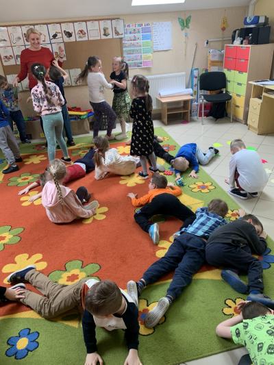 Dzieci w sali przedszkolnej bawią się w zabawę ,,Raz, dwa, trzy dinozaur patrzy. Nauczyciel stoi przed dziećmi a one stoją lub leżą na dywanie w przyjmując różne pozy