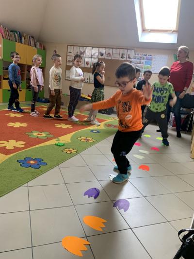 Dzieci w sali przedszkolnej ustawione są po kolei w długim rzędzie i skaczą w różnych kierunkach po śladach dinozaurów wyciętych z kolorowych kartek.