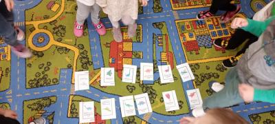 Dzieci ćwiczą na dywanie wykonując zadania z kart z dinozaurami rozłożonych na środku dywanu..jpg