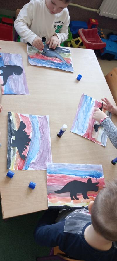 Dzieci wykonują pracę plastyczną przy stoliku, naklejając na kolorowym tle dinozaura