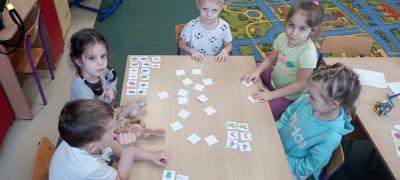 Grupa dzieci siedzi przy stoliku i grają w  dinozaurowe memory. Dzieci odkrywają pary takich samych obrazków z dinozaur