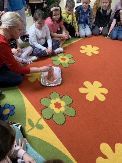 Nauczyciel prezentuje dzieciom zgromadzonym na dywanie w sali przedszkolnej jak wygląda wybuch wulkanu i wypłynięcie z niego lawy