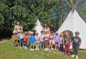 Grupa dzieci wraz z dwoma paniami pozuje do zdjęcia. Za nimi drzewa i 2 indiańskie namioty tipi. Grupa dzieci wraz z dwoma paniami pozuje do zdjęcia. Za nimi drzewa i 2 indiańskie namioty tipi.