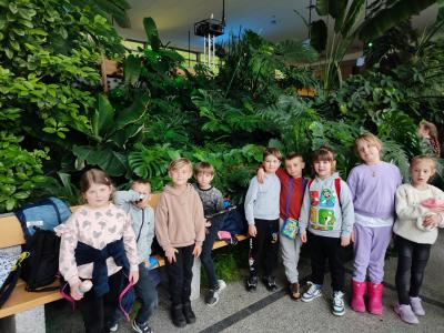 Dzieci stoją w rzędzie, za nimi zielone liście roślin
